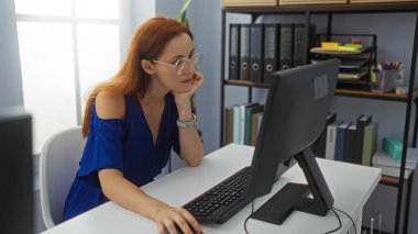 Ofiste bilgisayar başında çalışan, gözlüklü ve mavi elbiseli, raflar ve klasörlerle çevrili, düşünceli bir ifadeyle işine konsantre olan bir kadın..