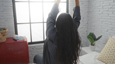 Yatakta oturan bir kadın geniş pencereli modern bir yatak odasında kollarını esnetiyor, uzun saçlarını ve rahat bir sabah rutini çiziyor..