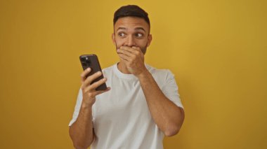Beyaz tişörtlü yakışıklı, sakallı, elinde akıllı bir telefon tutan, neşeli sarı bir arka planda izole edilmiş genç bir İspanyol..