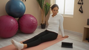 Woman stretching on yoga mat in gym with exercise balls and plants by window clipart