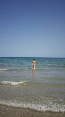 Kumsalda güneşli bir günün tadını çıkaran bir kadın durgun bir deniz ve mavi gökyüzü ufkuyla berrak suda duruyor.