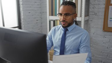 Ofis ortamında çalışan, bilgisayar ekranına odaklanmış, gözlük takan ve kravat takan, profesyonel bir ortamda çalışan genç bir adam..