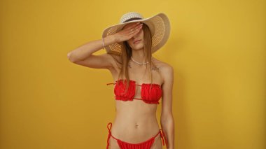 Kırmızı bikinili ve güneş şapkalı genç, çekici bir kadın sarı arka plan duvarına karşı poz veriyor, yüzünü eliyle kapatıyor, yaz havasını ve moda estetiğini somutlaştırıyor..