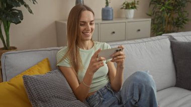 Genç, çekici sarışın bir kadın akıllı telefon kullanarak gülümsüyor, modern, rahat bir oturma odasında koltukta rahatça oturuyor..