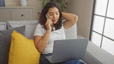 Genç, çekici, İspanyol bir kadın oturma odasında bilgisayarında çalışırken akıllı telefonuyla konuşuyor, rahat bir şekilde rahat bir koltukta canlı sarı bir yastıkla oturuyor..