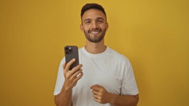 Yakışıklı, sakallı, sarı arka plan duvarına karşı akıllı telefon tutan, kendine güvenen ve gülümseyen bir İspanyol..