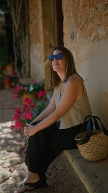 Güneşli bir günün tadını çıkaran bir kadın Mallorca 'da dışarıda oturuyor, İspanya' da bir terasta canlı çiçekler ve taşralı duvarlarla çevrili.