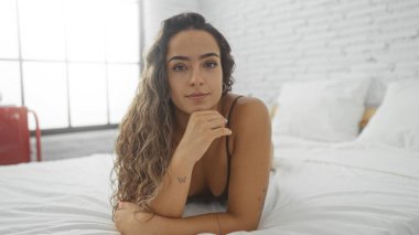 Genç, çekici, İspanyol bir kadın, parlak, minimalist bir yatak odasında uzanmış, esmer saçlarını ve dövmelerini sergiliyor, rahat ve kendinden emin bir ifadeyle.