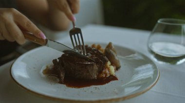 Zarif restoranda bıçak ve çatalla biftek kesen bir kadın, bir bardak su ve beyaz masa örtüsü atmosferiyle lezzetli bir yemek sunumuyla güzel bir yemek deneyimini ön plana çıkarıyor.