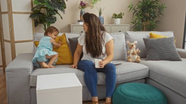 Kanepede oturmuş kahve içen bir kadın, sıcak ve parlak bir oturma odasında genç kızıyla etkileşime geçiyor, sevgi ve beraberlikle dolu sıcak bir aile anını gösteriyor.