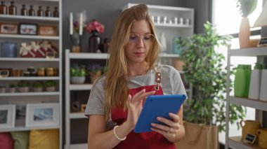 Genç, kadın, sarışın, dekoratif tablet kullanıyor, ev dekorasyonu dükkanı, renkli ürünler ve yeşillik raflarıyla çevrili.