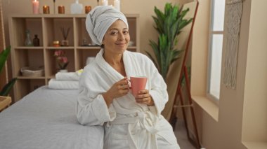 Bornoz ve havlu sarığı giymiş bir kadın, elinde mumlar ve bitkilerle dolu sıcak bir spa odasında elinde fincan tutarken gülümsüyor.