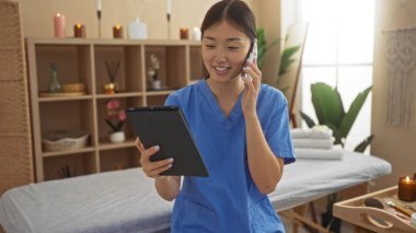 Kaplıca odasında elinde tabletle telefonla konuşan, mavi üniforma giyen, mumlar ve sağlık dekorasyonlarıyla çevrili bir kadın, sakin ve profesyonel bir atmosfer sağlıyor..