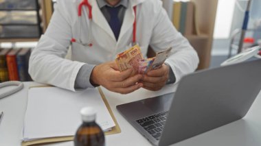 İspanyol bir erkek doktor klinikteki masasında Meksika pezosu sayıyor. Tıbbi ekipman ve belgeler etrafa saçılmış durumda..