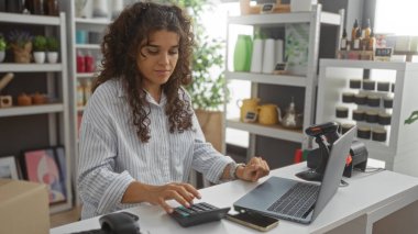 Genç, çekici, Latin kökenli, ev dekorasyonu ve modern iç dekorasyon ürünleriyle donatılmış bir iç dekorasyonda dizüstü bilgisayar ve hesap makinesiyle çalışan bir kadın.