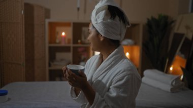 Sıcak bir sağlık merkezinde sıcak bir içecek içen, arka planda mumlarla lüks bir salonun huzurlu atmosferini yakalayan İspanyol bir kadın..