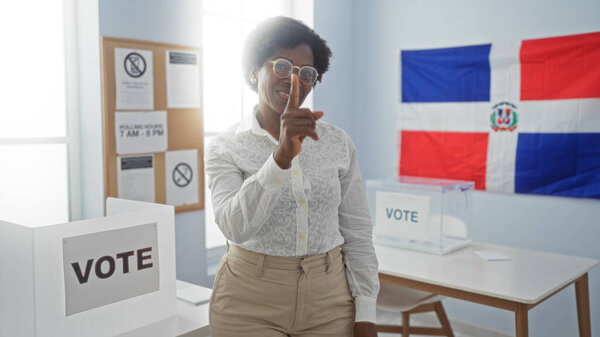 Африканская американка в помещении доминиканской республики избирательная комната с флагом и табличкой для голосования, указывающая пальцем в камеру, демонстрирующая участие в выборах.