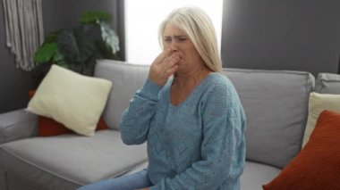 İçerideki kadın, kötü kokudan dolayı burnu kapalı, parlak, modern oturma odasındaki koltukta oturuyor. Etrafında yastıklar ve arka plandaki bitkiler rahatsız edici.