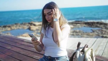 Telefon kullanan bir kadın açık deniz kenarında geziniyor, saati kontrol ediyor, yaz ve okyanus manzarasını ahşap bankta sırt çantasıyla sergiliyor..