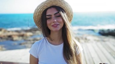 Hasır şapka takan genç bir kadın arka planda okyanusla birlikte parlak güneşin altında güzellik ve kendine güven yayıyor..