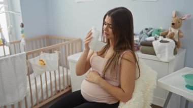 Bebek odasında oturan hamile bir kadın beşikteki dokuyla duygusal hissediyor oyuncaklarla çevrili annelik beklentisi ve hazırlığını ifade ediyor..