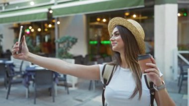 Şehir sokaklarında selfie çeken bir kadın elinde şapka ve sırt çantasıyla elinde kahveyle şehirde oturma düzeniyle gün boyunca çekici bir ifade ve esmer saçla geziyor.