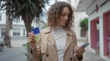 Şehirde elinde kredi kartı ve akıllı telefonla gülümseyen kıvırcık saçlı ve kahverengi ceketli bir kadın, modern şehir yaşam tarzını ve alışveriş kolaylığını temsil ediyor..