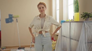 Oturma odasını silindirle boyayan kadın, yenileme projesi sırasında arka planda aletlerle yeni ev içinde gülümsüyor.