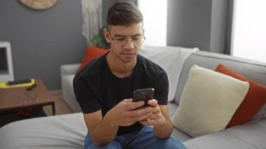 Genç adam rahat bir evde, yastıklı ve modern dekorlu akıllı telefon kullanıyor. Rahat ve birbirine bağlı bir atmosferi içeri taşıyor..