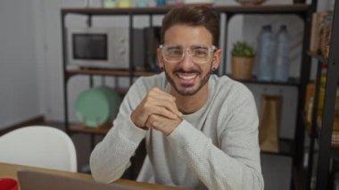 İspanyol bir adam çalışma odasında modern dekorla gülümsüyor, gözlük ve süveter takıyor, profesyonel ve yaklaşılabilir bir tavır sergiliyor..