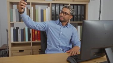 Sakallı ve gözlüklü genç adam dosya ve kitaplarla dolu bir ofiste selfie çekiyor, modern bir iş yerinde rahatça oturuyor.