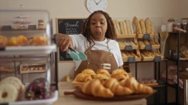 Fırında çalışan, ekmek ve hamur işleri yapan, önlük takan, maşa tutan, fırında pişirilmiş mallarla çevrili bir kadın, yoğun bir dükkan ortamı sergiliyor..