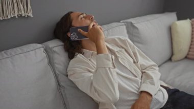 Modern bir oturma odasındaki kanepede otururken akıllı telefondan konuşan uzun saçlı genç adam.