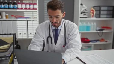 Tıbbi malzemelerle çevrili bir klinikte dizüstü bilgisayarda çalışan İspanyol erkek doktor iyi organize edilmiş bir hastane ortamında odaklanmış ve profesyonel görünüyor..