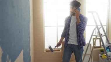 Duvarı boyayan genç adam telefonda, evde, ekose gömlek giyiyor, merdivenle ayarlı ve rahat oturma odasında doğal ışık var..