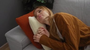 Kanepede oturan, kahverengi kazak giyen, rahat bir evde yastık tutan, modern bir oturma odasında huzur ve sükunet sergileyen bir kadın..