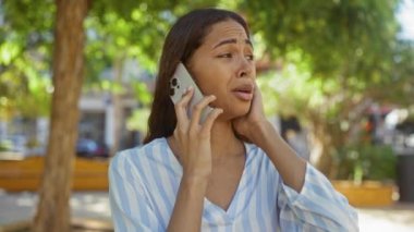 Telefonda konuşan genç bir kadın, arka planda ağaçlar ve banklar olan bir şehir parkında endişeli görünüyor.