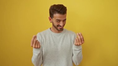 Süveter giyen genç İspanyol adam, kendine güvenen bir gülümseme ile mizah ve oyun ifade eden, canlı sarı bir arka plana karşı para hareketi yapıyor..
