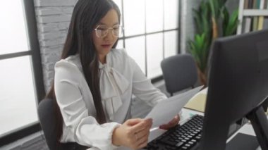 Parlak ofis ortamında bilgisayar ve bitkilerle, odaklanmış ve profesyonel atmosferle, gözlüklü ve zarif beyaz bluzlu bir kadın evrak okuyor..