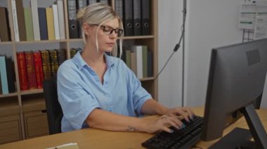 Ofiste çalışan bir kadın, sarışın, çekici, gözlüklü, bilgisayarda daktilo kullanan, arka planda dosyaları olan, çalışma ortamı olan bir kadın