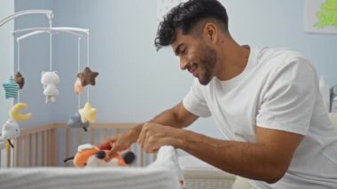 Genç adam, bebek oyuncağıyla rahat yatak odasında etkileşime giriyor. Ev ortamındaki sıcaklık ve neşeyi yansıtıyor. Ebeveyn ve çocuk arasında hassas bir an yaratıyor..