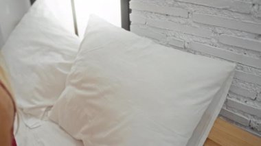 Evin içinde yatakta dinlenen, gözleri kapalı gülümseyen, sarı saçlı, kırmızı bluzlu, huzurlu bir yatak odası ortamında beyaz tuğla bir duvara yaslanmış bir kadın..