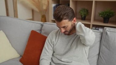 Oturma odasındaki rahat bir kanepede oturan boynu ağrıyan genç adam. Etrafı minderler ve bitkilerle çevrili. Rahatsızlık ve rahatlama ihtiyacını iletiyor..
