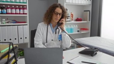 Klinikteki kadın doktor telefonla konuşuyor, bilgisayarla ve belgelerle çalışıyor, tıbbi malzemeler ve kitaplarla çevrili.