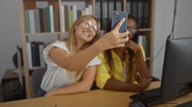 Ofis ortamında selfie çeken ve gülümseyen kadınlar, dost canlısı bir iş ortamı sergiliyorlar.