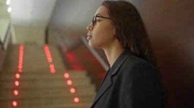 Siyah ceketli genç bir kadın. Kapalı bir lobide kırmızı ışıkları olan merdivenlerin yanında duruyor.
