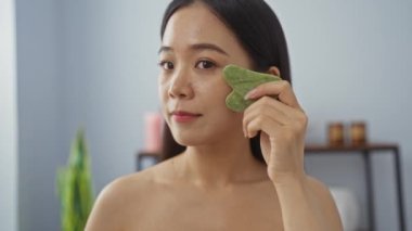 Genç bir kadın spa odasında yeşim bir aletle yüz masajı yapıyor. Rahatlamış bir ortamda sağlığın ve cildin önemini vurguluyor.