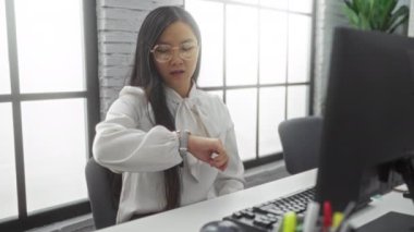 Modern ofisin içinde beyaz bluzlu bir iş kadını saati kontrol ediyor. Bilgisayarda çalışırken rahatsız görünüyor..