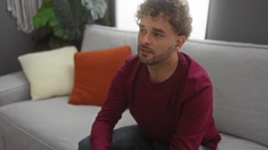 Kıvırcık saçlı genç adam rahat bir oturma odasındaki kanepede sıkıntılı bir şekilde oturuyor, elleri yüzlerinde, ev ortamında duygularını ifade ediyor..