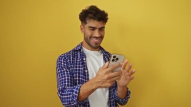 Kareli tişörtlü İspanyol adam, açık sarı arka planda video görüşmesinde gülümsüyor..